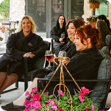 - The Renaissance Day Spa & Alternative Wellness, Fayetteville NC Four women in black robes sit on outdoor lounge chairs, smiling and holding glasses of wine, with pink flowers in the foreground—enjoying a relaxing moment after indulging in luxury day spa packages. - The Renaissance Day Spa & Alternative Wellness, Fayetteville NC