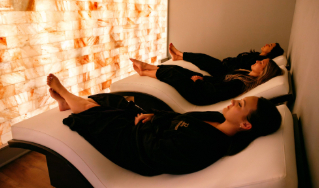 - The Renaissance Day Spa & Alternative Wellness, Fayetteville NC Three people in black robes lie on white reclining lounge chairs in a dimly lit room with a glowing salt wall. - The Renaissance Day Spa & Alternative Wellness, Fayetteville NC