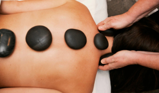 - The Renaissance Day Spa & Alternative Wellness, Fayetteville NC Person receiving a hot stone massage, with smooth black stones placed along their back and a therapist’s hands applying a stone to the shoulder area. - The Renaissance Day Spa & Alternative Wellness, Fayetteville NC