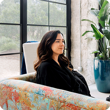 - The Renaissance Day Spa & Alternative Wellness, Fayetteville NC A woman with long dark hair sits on a colorful patterned sofa near a large window and a tall potted plant, enjoying the tranquil ambiance typical of luxury day spa packages. - The Renaissance Day Spa & Alternative Wellness, Fayetteville NC