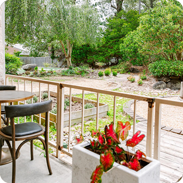 - The Renaissance Day Spa & Alternative Wellness, Fayetteville NC A patio with a table and chairs overlooks a garden with raised beds, gravel paths, lush greenery, and a planter with red flowers in the foreground—ideal for unwinding after luxury day spa packages. - The Renaissance Day Spa & Alternative Wellness, Fayetteville NC