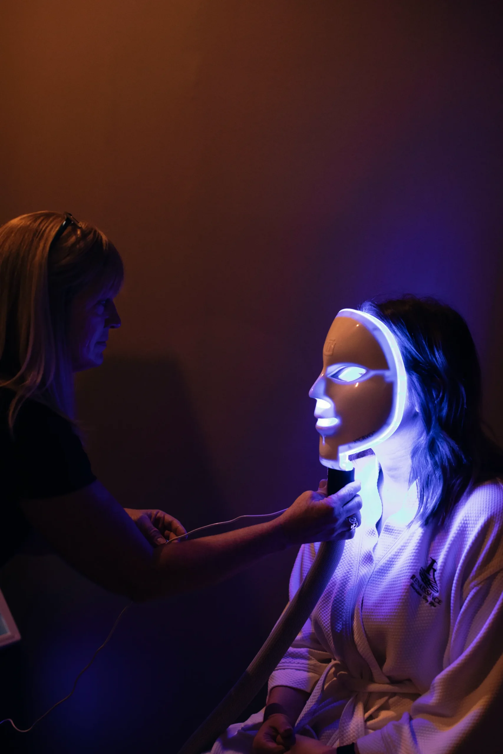 - The Renaissance Day Spa & Alternative Wellness, Fayetteville NC A person sits wearing an illuminated LED facial mask while another person adjusts a device under their chin in a dimly lit room. - The Renaissance Day Spa & Alternative Wellness, Fayetteville NC