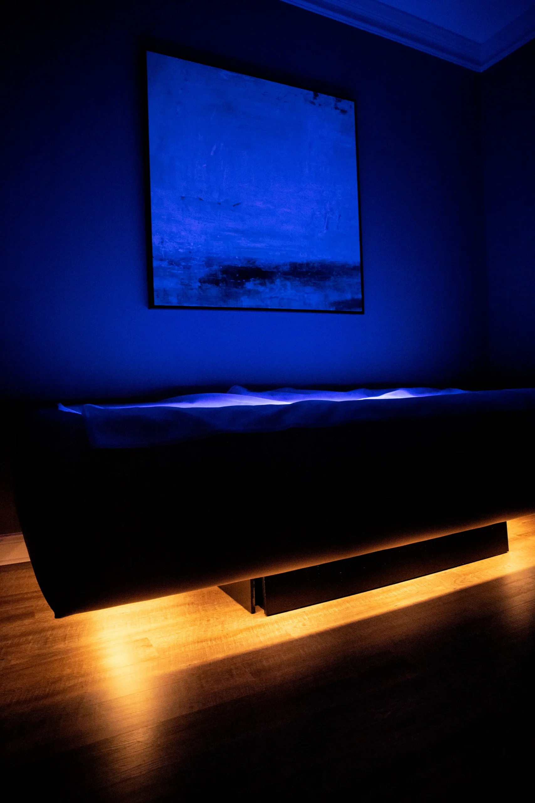 - The Renaissance Day Spa & Alternative Wellness, Fayetteville NC A dimly lit room with a glowing massage table, blue-toned artwork on the wall, and soft yellow lighting under the table on a wooden floor. - The Renaissance Day Spa & Alternative Wellness, Fayetteville NC